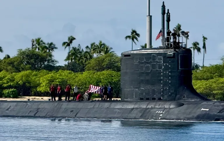 US Navy submarine surfaces near tropical island with crew on deck waving flag.