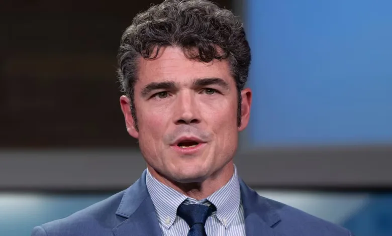 Man in blue suit and striped shirt looking at camera with serious expression. Curly dark hair.