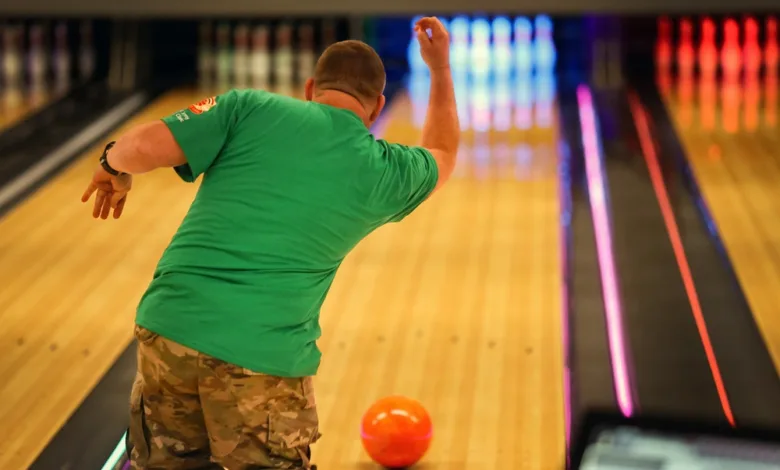 Man bowling with orange ball, green shirt and camo pants at bowling alley.