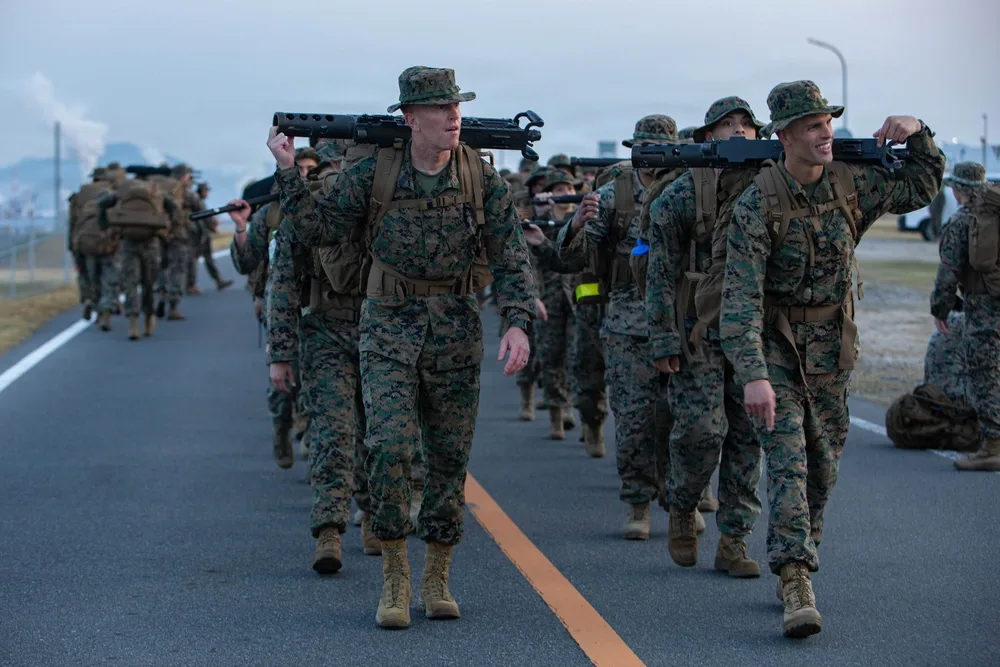 Marines hike with equipment at MCAS Iwakuni, wearing camouflage and carrying gear.