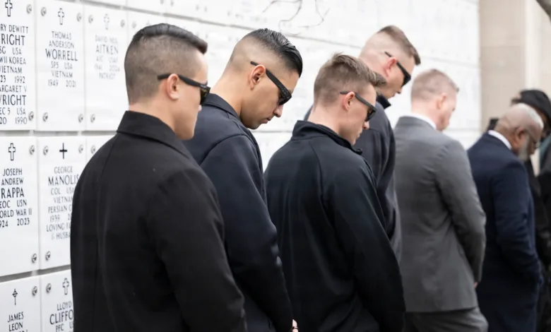 Men in dark suits pay respects at a memorial wall with names.
