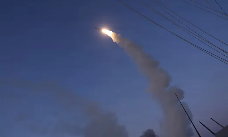 Missile launch with bright flame and smoke trail against a blue sky, ship visible in foreground.