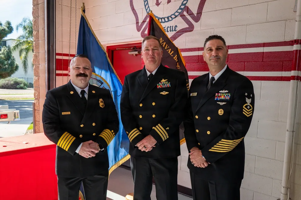 Three naval officers in uniform stand in front of a fire station backdrop.