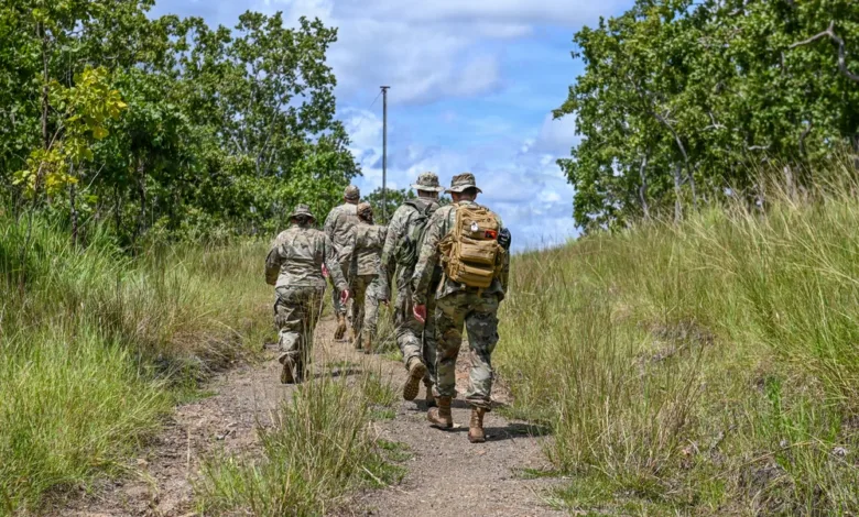 Soldiers hike on trail through grassy field, wearing camouflage and carrying gear.