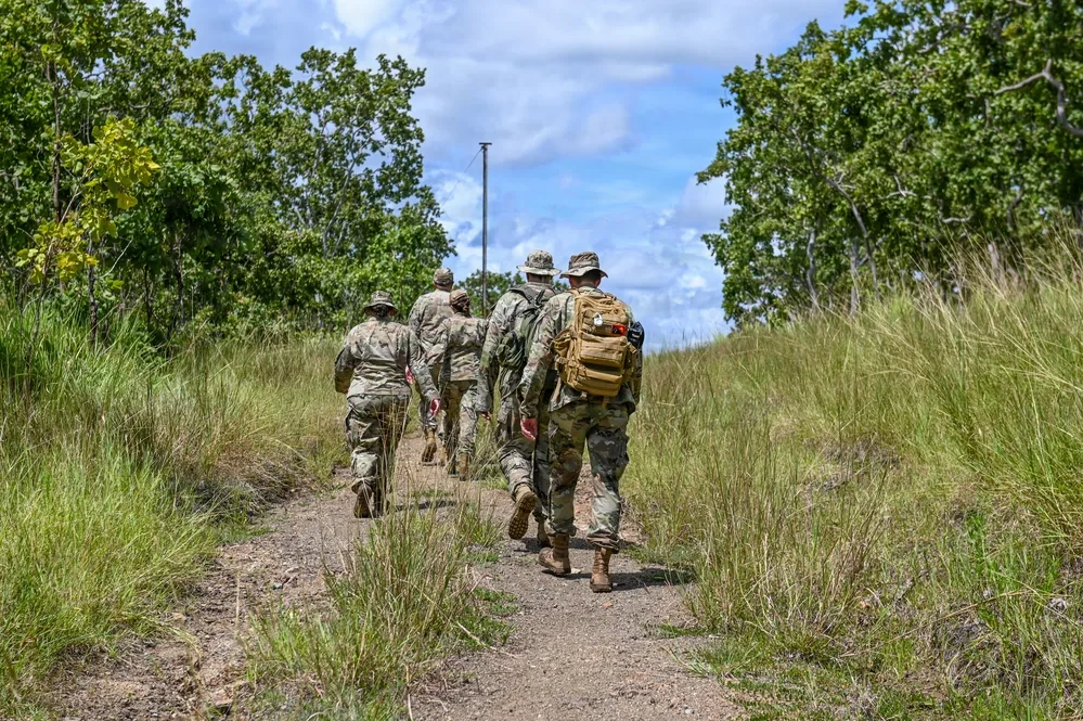 Soldiers hike on trail through grassy field, wearing camouflage and carrying gear.