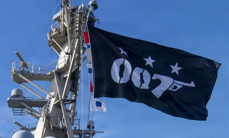 Black 007 flag with gun hoisted on ship mast against blue sky.