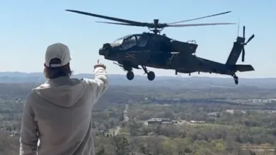 Man pointing at Apache helicopter in flight over wooded landscape.