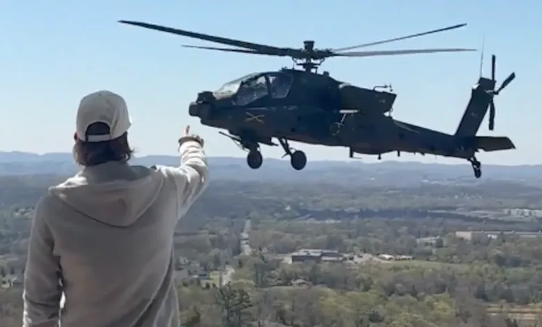Man pointing at Apache helicopter in flight over wooded landscape.