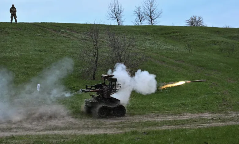 Armed ground robot fires projectile, kicking up smoke and dust on grassy hillside.