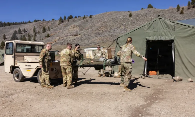 Army medics transport patient on stretcher to tent hospital. US Army vehicle visible.