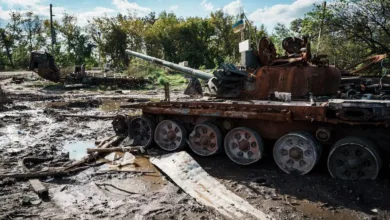 Destroyed Russian tank in Ukraine, muddy field, Ukrainian flag visible in background.