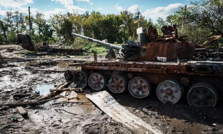Destroyed Russian tank in Ukraine, muddy field, Ukrainian flag visible in background.