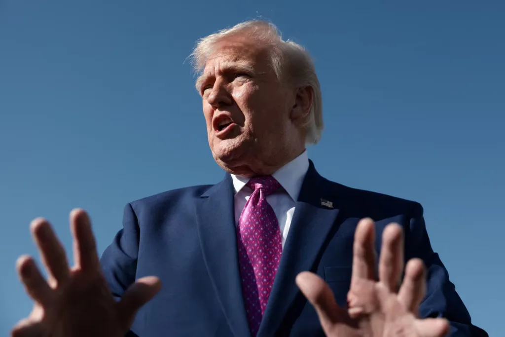 Donald Trump gestures with hands, wearing suit and tie against blue sky.