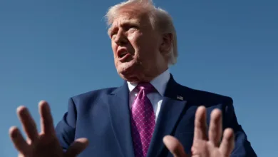 Donald Trump gestures with hands, wearing suit and tie against blue sky.
