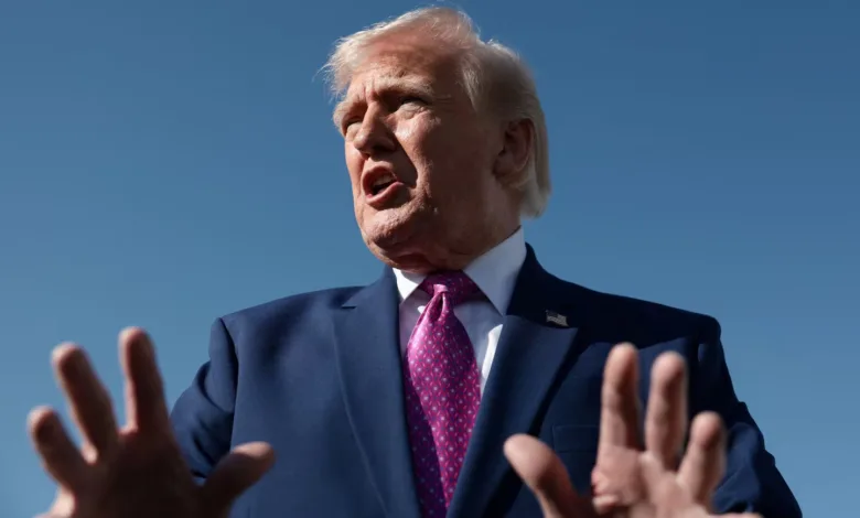 Donald Trump gestures with hands, wearing suit and tie against blue sky.