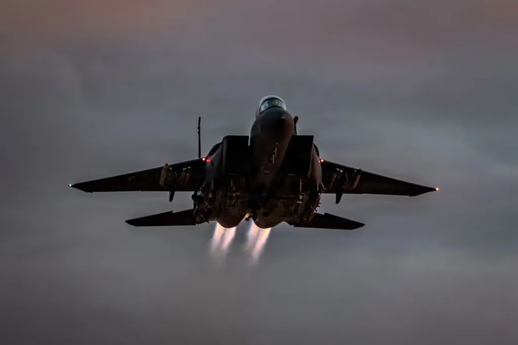 F-15 Eagle fighter jet taking off with afterburners engaged against a cloudy sky.