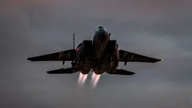 F-15 Eagle fighter jet taking off with afterburners engaged against a cloudy sky.