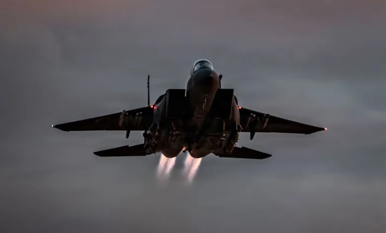 F-15 Eagle fighter jet taking off with afterburners engaged against a cloudy sky.