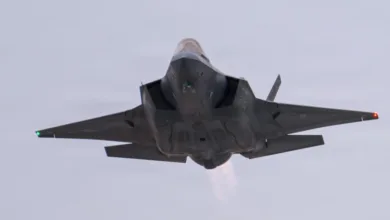 F-35 fighter jet flying, viewed from below. Gray aircraft with engine exhaust visible against a light sky.