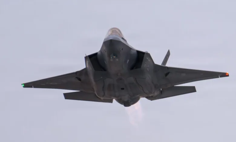 F-35 fighter jet flying, viewed from below. Gray aircraft with engine exhaust visible against a light sky.