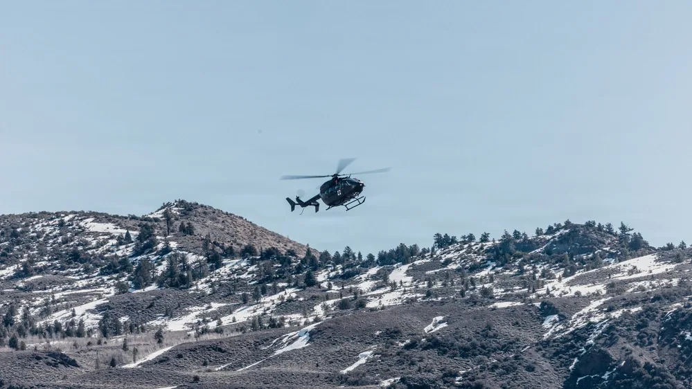 Black helicopter flying over snowy mountains on a clear day.