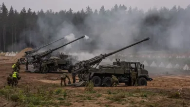 Military howitzers firing during an exercise with soldiers nearby. Smoke is visible.