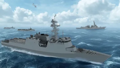 Modern naval frigates and cargo ships sail in formation on the ocean under a cloudy sky.