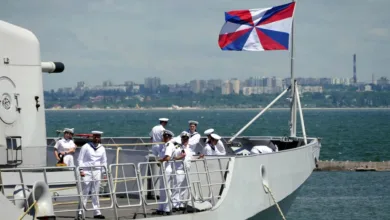 Naval officers on ship deck under naval flag, city skyline background.