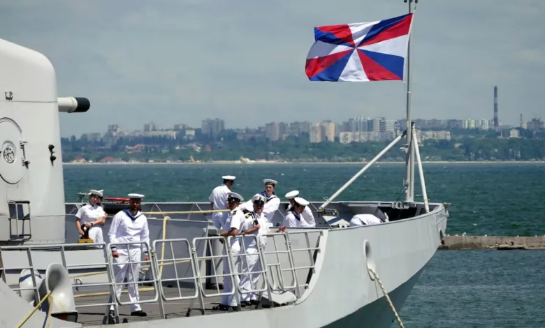 Naval officers on ship deck under naval flag, city skyline background.