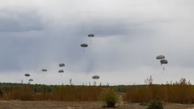 Paratroopers descending with parachutes over a wooded area against a cloudy sky.