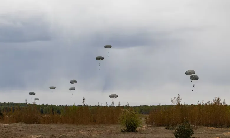 Paratroopers descending with parachutes over a wooded area against a cloudy sky.