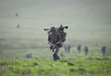 Soldier with backpack and rifle walks across a grassy field during military training exercise.