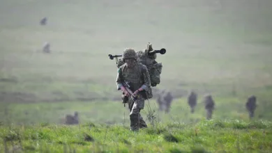 Soldier with backpack and rifle walks across a grassy field during military training exercise.