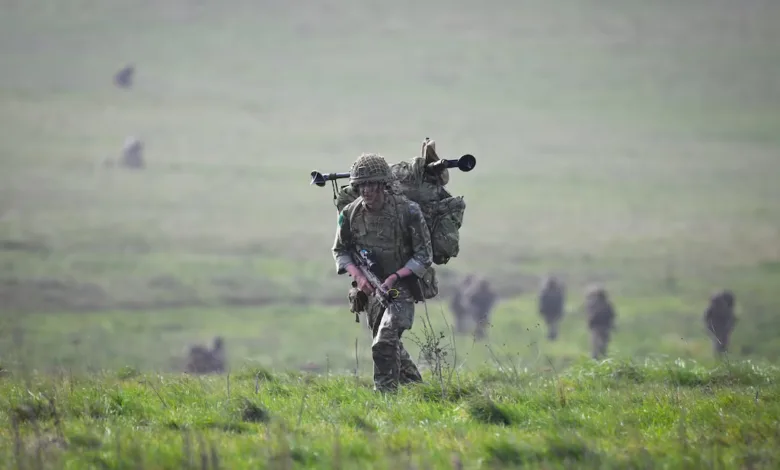 Soldier with backpack and rifle walks across a grassy field during military training exercise.