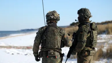 Two soldiers in camouflage gear stand on a snowy beach, looking out at the ocean.