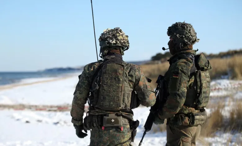 Two soldiers in camouflage gear stand on a snowy beach, looking out at the ocean.
