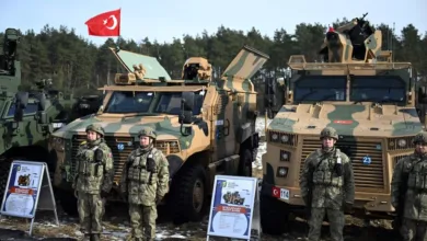 Turkish soldiers stand before armored vehicles with Turkish flag in background.