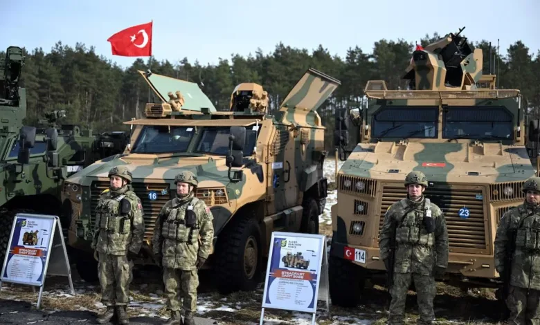 Turkish soldiers stand before armored vehicles with Turkish flag in background.