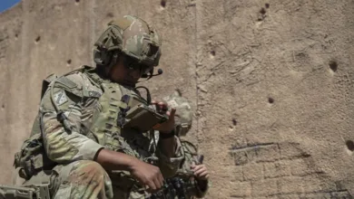 U.S. Army soldier in tactical gear adjusts equipment near a wall.