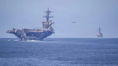 US Navy aircraft carrier and destroyer sailing on the ocean with helicopter overhead.