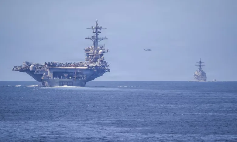 US Navy aircraft carrier and destroyer sailing on the ocean with helicopter overhead.