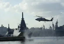 Naval helicopter flies over a large warship in a harbor with other ships docked.