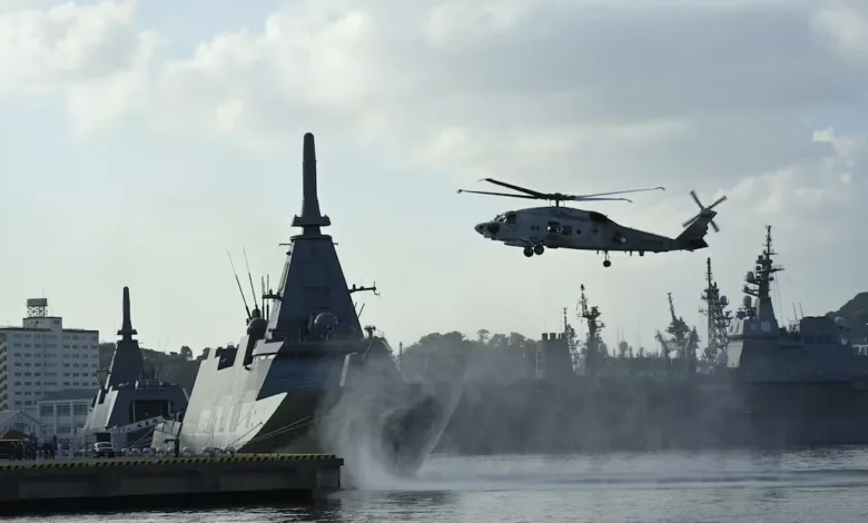 Naval helicopter flies over a large warship in a harbor with other ships docked.