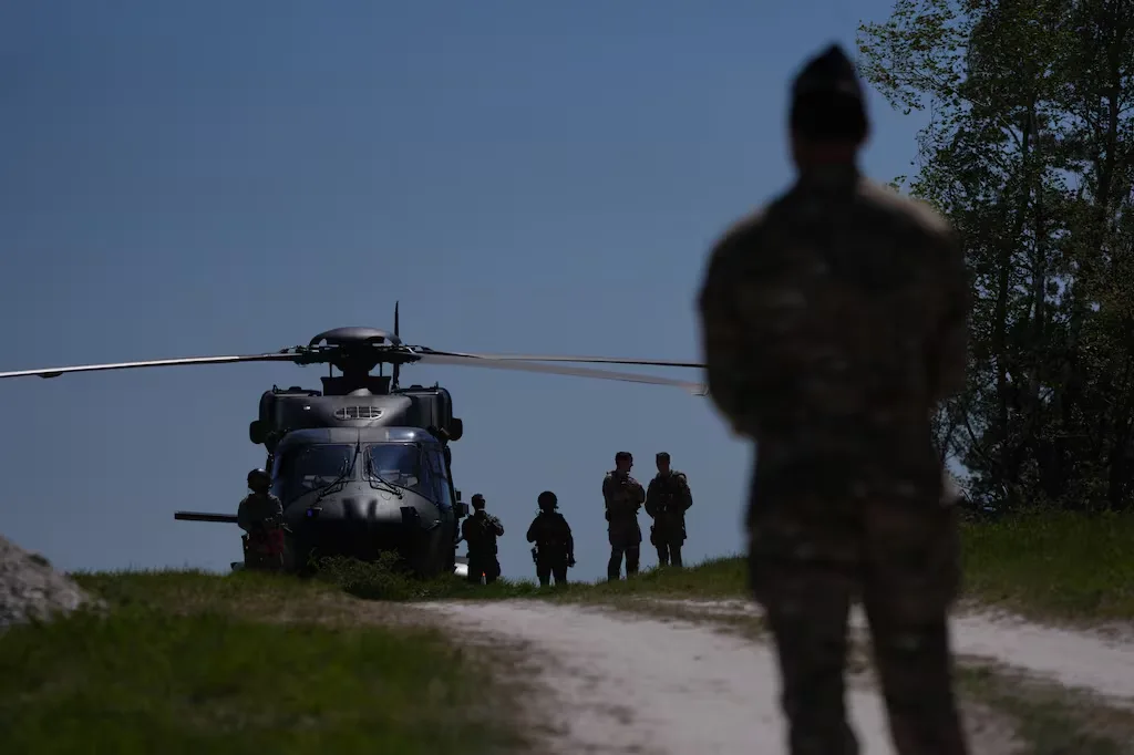 Military personnel stand near a Black Hawk helicopter on a grassy field.