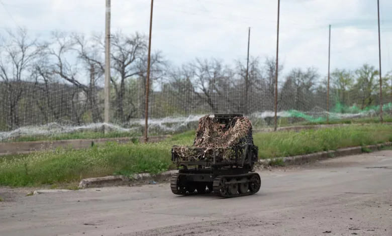 Camouflaged tracked robot moves on a paved road with trees and sky in the background.