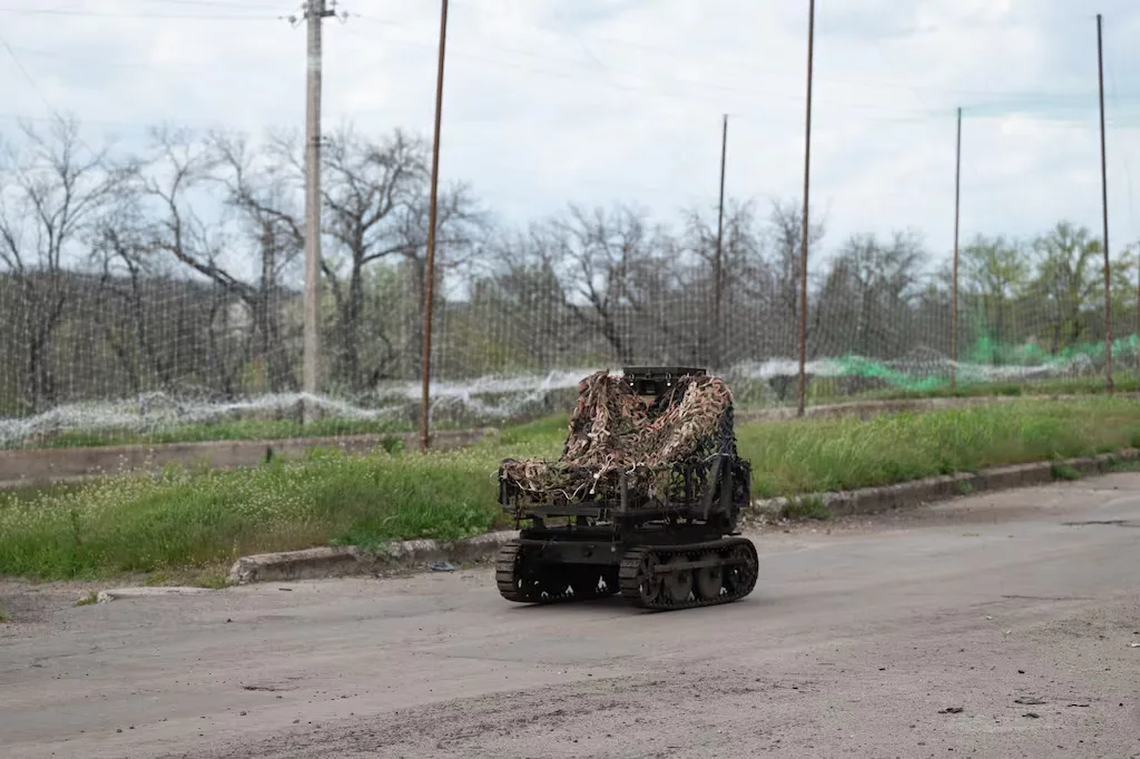 Camouflaged tracked robot moves on a paved road with trees and sky in the background.