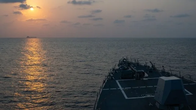 Naval destroyer sails at sunset with cargo ship on horizon.