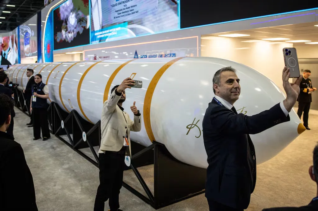 Men take photos of a large white rocket stage with gold stripes at an exhibition.
