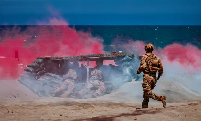 Soldiers train with smoke grenades on a beach near a fortified position.