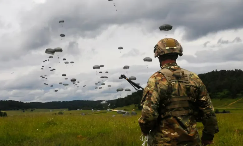 Soldiers parachute onto a grassy field with many parachutes descending.